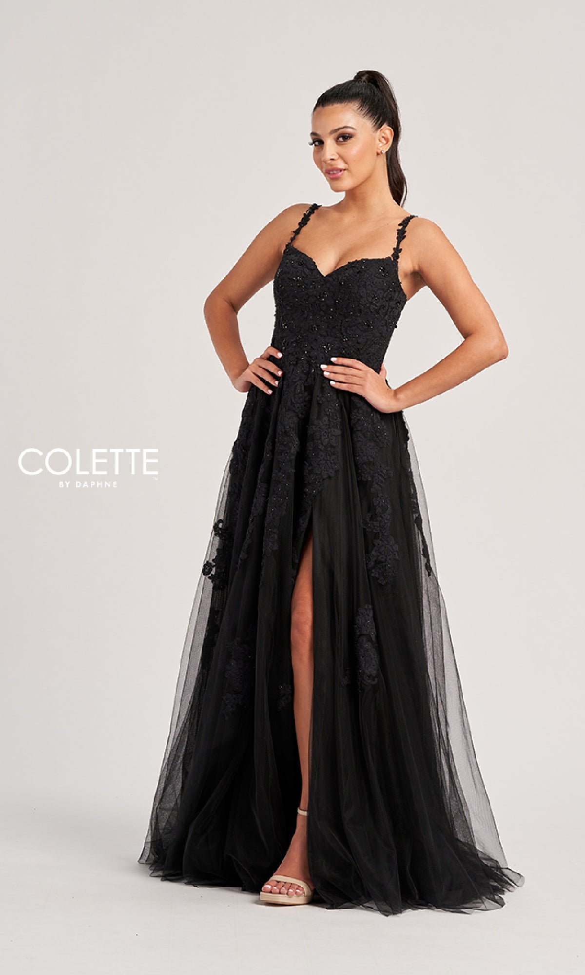 A woman stands against a plain light background wearing the Colette CL8220 long prom dress—a sleeveless black evening gown with floral detailing, a front slit, and an elegant lace-up back.